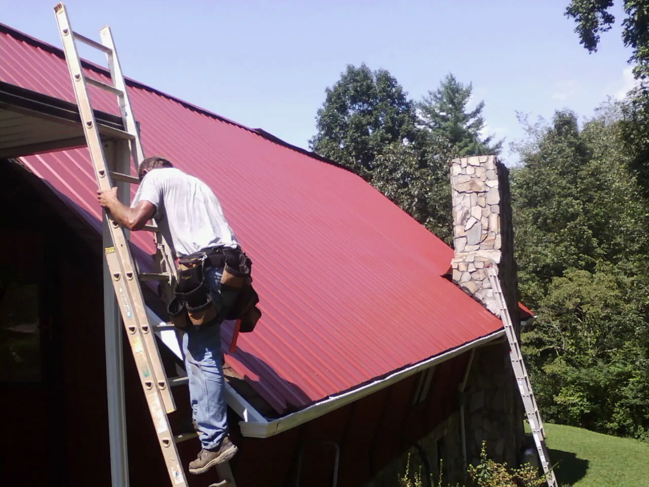 Roofer on a ladder beside a red metal roof and stone chimney — Soffit & Fascia Repair by Roofing Brothers in Northridge